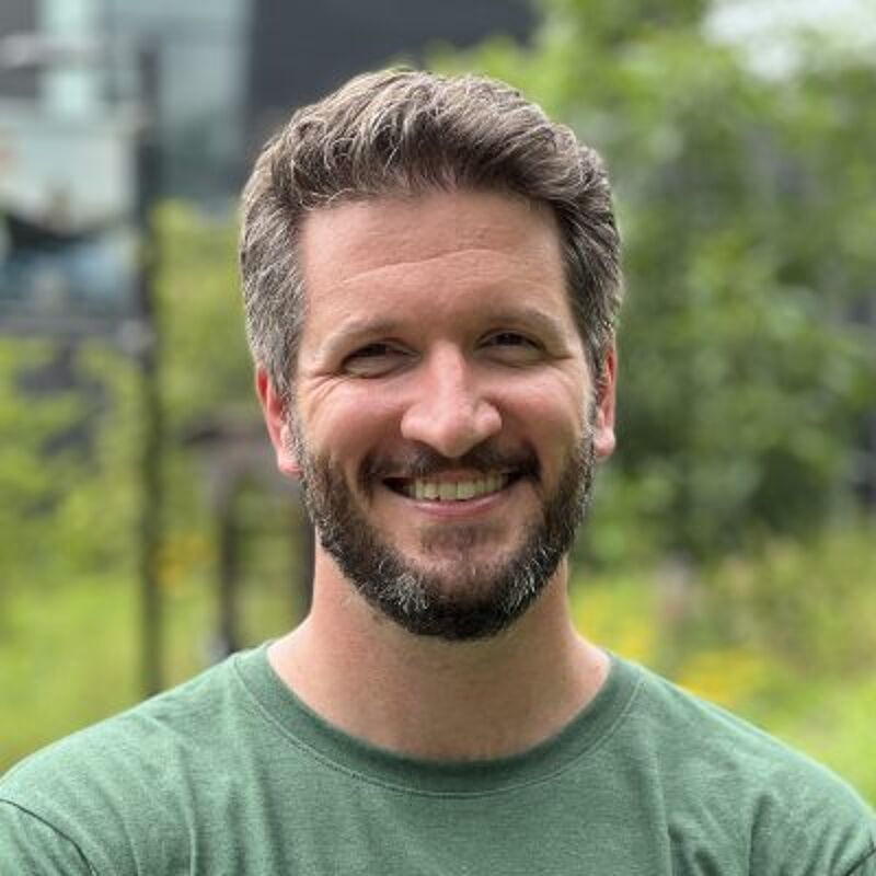 The image shows a man with short, graying hair and a beard. He is wearing a green t-shirt and smiling at the camera. The background is blurred, but it appears to be outdoors with trees and greenery. The man seems to be in a park or a similar natural setting.